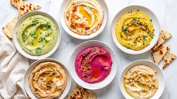 Seis tazones de hummus de distintos colores con aceite, especias, hierbas, garbanzos, semillas. Pan pita y un paño blanco sobre una superficie de mármol.