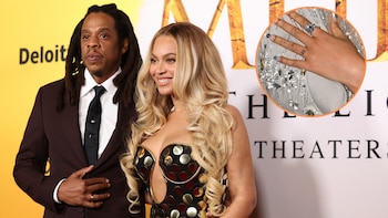 Jay-Z en traje oscuro y Beyoncé en vestido negro y dorado con recortes, cabello rubio, posan. Detalle circular de la mano de Beyoncé con un anillo