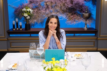 Juana Viale, con cabello oscuro y camisa azul claro, sonríe con las manos bajo la barbilla, sentada en una mesa blanca con copas y pan