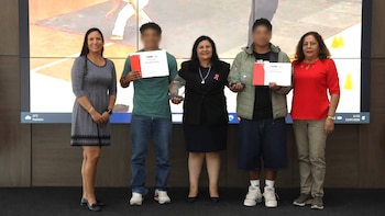 Cinco personas de pie frente a una pantalla digital grande; tres mujeres y dos hombres jóvenes con los rostros borrosos sostienen premios y certificados