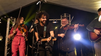 Una mujer vestida de rosa y un hombre con gafas tocan el violín junto a un guitarrista sobre un escenario con luces