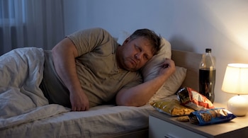 Hombre de mediana edad con obesidad, acostado en la cama con expresión de insomnio. Junto a él, una mesita de noche con botellas de bebida y paquetes de comida.