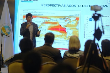 Hombre con micrófono y puntero láser frente a pantalla con mapa de temperaturas oceánicas y gráficos, con audiencia, bandera de INSIVUMEH y trípode