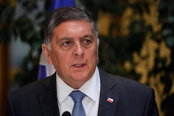 Un hombre mayor de traje oscuro, camisa blanca y corbata azul habla con un prendedor de la bandera de Chile en la solapa