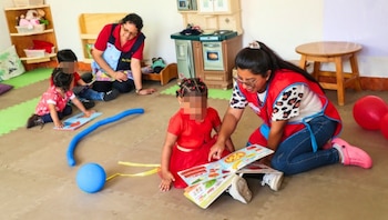 Dos mujeres leen libros ilustrados con niños en una sala de juegos equipada con una cocinita de juguete y estantes