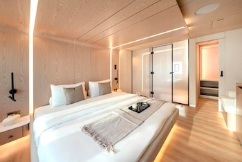 Habitación con cama matrimonial vestida de blanco, paredes y techo revestidos en paneles de madera clara, lámparas y puerta de vidrio
