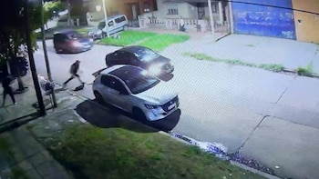 Cámara de seguridad nocturna en calle residencial: un coche subcompacto plateado y vehículos oscuros; varias personas se desplazan por la acera