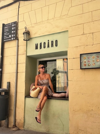 Una mujer rubia con vestido estampado, sandalias y lentes de sol, sentada en el alféizar de una ventana de un local Mocàro. Fondo: pared amarilla y cartel