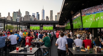 Vista aérea de personas en un bar al aire libre viendo partidos de fútbol en pantallas gigantes, con el horizonte de la ciudad de Nueva York al fondo.