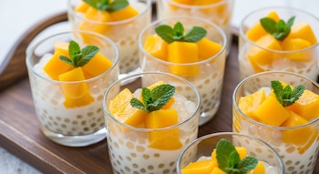 Vista de cerca de múltiples vasitos de postre, cada uno con perlas de tapioca, una capa blanca, cubos de mango y una ramita de menta.