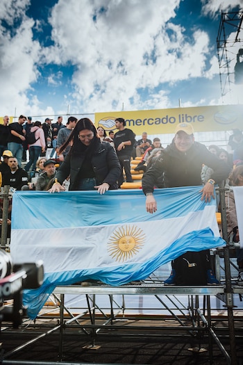 Dos personas sostienen una bandera argentina en las gradas de un evento al aire libre con un cartel de Mercado Libre al fondo y cielo azul con nubes
