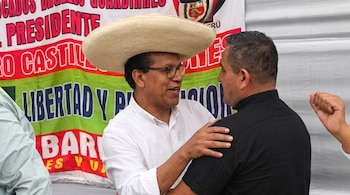Primer plano de dos hombres conversando. Uno lleva sombrero de paja y gafas, el otro está de espaldas con camisa negra. Un banner peruano detrás.
