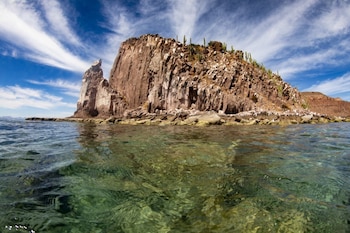 Un islote rocoso cubierto de cactus emerge de aguas cristalinas que muestran rocas sumergidas, bajo un cielo azul con nubes blancas y alargadas