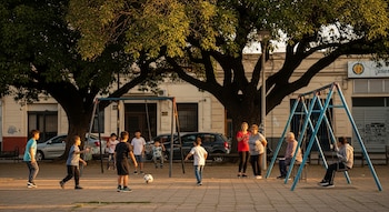 Seis niños juegan con una pelota, otro niño en una hamaca, dos adultos de pie y un adulto mayor sentado en un columpio en una plaza arbolada.