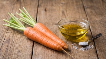 Dos zanahorias con hojas verdes, un recipiente de vidrio con aceite y una cuchara con sal sobre una mesa de madera.