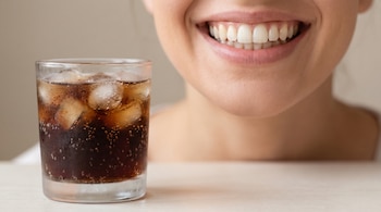 Primer plano de boca sonriendo con dientes blancos y encías. Un vaso con bebida gaseosa oscura y cubos de hielo está desenfocado en primer plano.