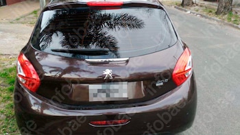 Parte trasera de un automóvil Peugeot 208 color oscuro estacionado en la calle. El vehículo tiene luces traseras rojas y reflejos de palmeras en el cristal