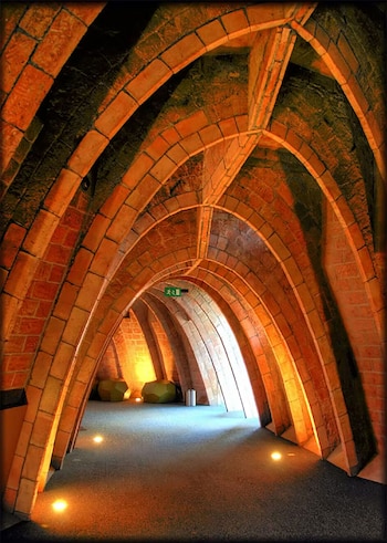 Un pasillo con múltiples arcos catenarios de ladrillo visto, iluminado desde el suelo, crea una perspectiva profunda. Al fondo, una salida luminosa y dos elementos orgánicos verdes