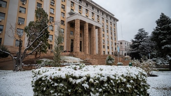 Fachada de un edificio beige con columnas de piedra en la entrada, escaleras, árboles y arbustos con nieve sobre el suelo y un cielo nublado