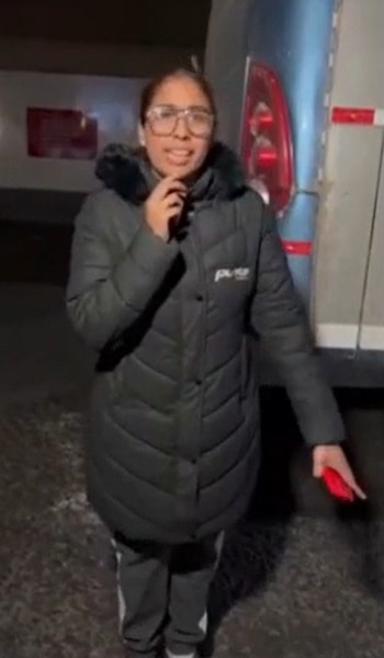 Mujer joven con gafas y abrigo oscuro de invierno, de pie al aire libre, con un autobús o vehículo grande y una barrera al fondo