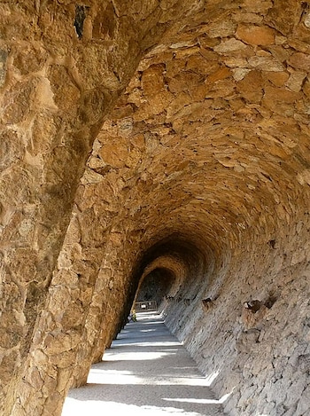 Pasaje curvo de piedra natural en el Park Güell, con un techo abovedado y pilares inclinados que crean un efecto serpenteante. La luz solar ilumina el suelo