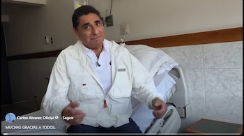 Carlos Álvarez, sentado en una cama de hospital, viste una chaqueta blanca y polo, gesticulando hacia la cámara en una habitación con paredes claras