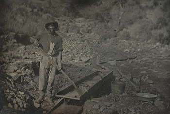 Hombre de pie con sombrero y pala junto a una estructura de madera para lavar mineral en un terreno rocoso
