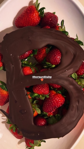 Primer plano de una bandeja blanca llena de fresas rojas frescas con tallos verdes, cubierta parcialmente por una gran letra 'B' de chocolate oscuro