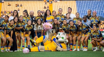 Veinticuatro personas y una mascota de tigre con uniformes de fútbol amarillos y azules posan en un campo con una pelota de fútbol y un ramo de flores.