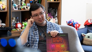 Hombre con gafas y camisa de cuadros azul y blanca sostiene un libro ilustrado. Al fondo, estantería con figuras coleccionables de videojuegos y un sofá azul