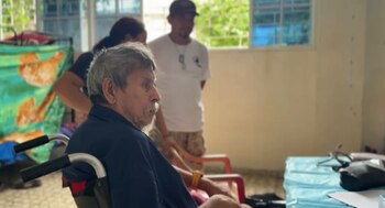 Un adulto mayor permanece en un albergue temporal, acompañado por personal de atención y familiares, en el contexto de las recientes condiciones climáticas (Cortesía Noticiero El Salvador).