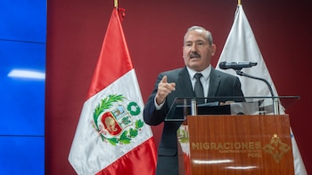 Un hombre en traje formal habla desde un podio de Migraciones Perú, flanqueado por la bandera peruana y un micrófono