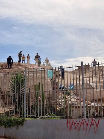 Varias personas de pie sobre una huaca rocosa con basura, detrás de una valla metálica con pinchos. Abajo, plantas y una pared con graffiti rojo