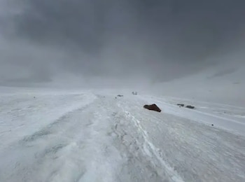 Amplio paisaje invernal con terreno cubierto de nieve, huellas en la superficie, rocas dispersas y un cielo denso con nubes grises