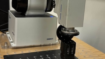 Brazo robótico de color blanco y gris con un sensor negro ubicado sobre una placa horizontal con signos en braille
