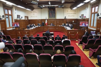 Sala de audiencias con asientos rojos, mesa de madera, magistrados, público, pantallas, bandera de México, retrato y textos en las paredes