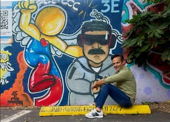 Boris Peraza sonríe, sentado en un bordillo de estacionamiento amarillo con la inscripción "PAID PARKING 24/7" y un mural de graffiti colorido detrás que incluye dos figuras