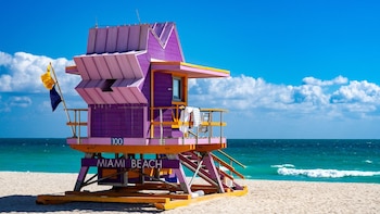 Caseta de salvavidas morada y rosa con la inscripción Miami Beach sobre la arena, frente al mar azul y bajo un cielo con nubes