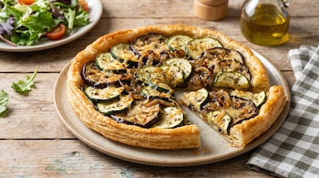 Una tarta de berenjena y zucchini con corteza dorada y una porción ya servida se muestra en un plato sobre una mesa de madera, con una ensalada al fondo.