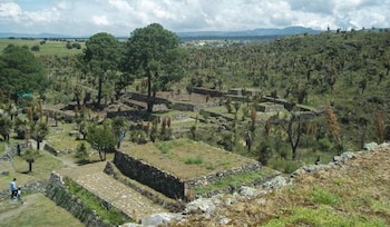 Estructuras de piedra, muros, plataformas, caminos empedrados, árboles, arbustos, vegetación, montañas al fondo, cielo nublado, dos personas