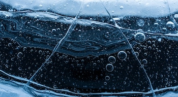 Plano detalle de petróleo líquido oscuro con burbujas de aire y vetas visibles, atrapado bajo una gruesa capa de hielo azulado y translúcido.