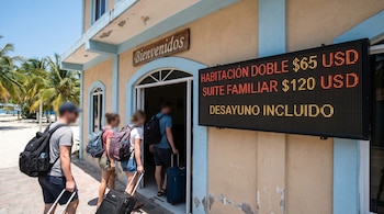 Tres viajeros con mochilas y maletas ingresan a un edificio. Un letrero 'Bienvenidos' y una pantalla digital con precios de hotel son visibles. Palmeras al fondo.