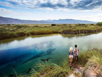 Vista superior de una pareja de espaldas al borde de un cuerpo de agua con tonos azules y turquesas. El entorno está cubierto de hierba, con montañas al fondo bajo un cielo nublado
