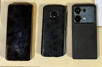 Tres teléfonos inteligentes de diferentes modelos, dos negros y uno con bordes dorados, yacen sobre una superficie clara, mostrando sus pantallas y cámaras traseras