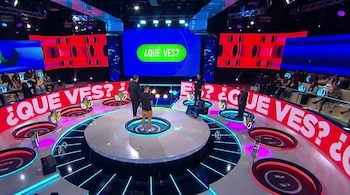 Escenario de televisión con plataforma circular, tres hombres, público sentado, pantalla con "¿QUÉ VES?" y paneles decorativos rojos.