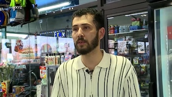 Hombre barbudo con camisa polo de rayas blancas y negras en una tienda, con estantes de productos y una puerta de vidrio al fondo