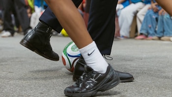 Piernas y pies pateando un balón de fútbol multicolor en una calle. Un pie lleva bota negra, el otro calcetín blanco Nike y zapatilla deportiva oscura
