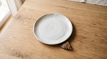 Un plato blanco de cerámica sobre una mesa de madera clara con la punta de una hoja seca debajo del borde.