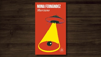 Primer plano de la portada del libro 'Marciano' de Nona Fernández, con un platillo volador rojo, un haz de luz amarillo y un círculo negro, sobre una superficie de madera oscura