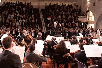Un director de orquesta dirige a una orquesta con instrumentos de cuerda frente a un público numeroso en un auditorio con asientos escalonados
